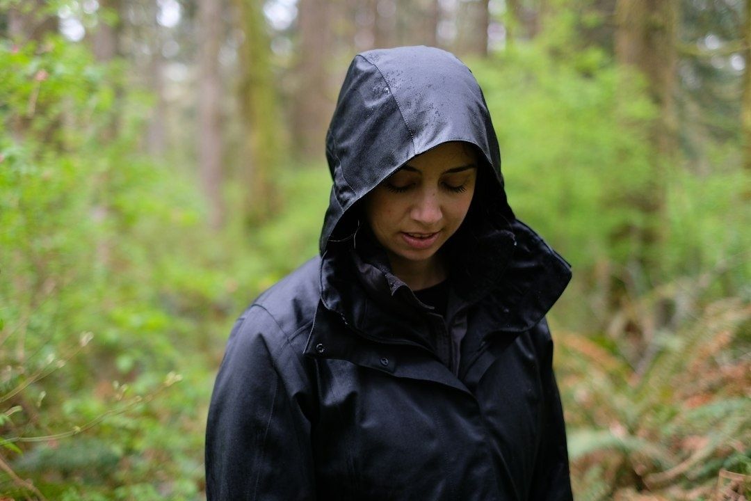 Person i svart huvad regnjacka står i en grönskande skog